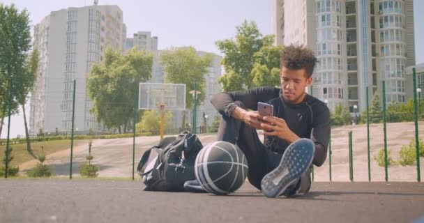 Closeup retrato de jovem afro-americano bonito jogador de basquete masculino ouvindo música em vibrações em seu telefone sentado com uma bola ao ar livre na quadra — Vídeo de Stock