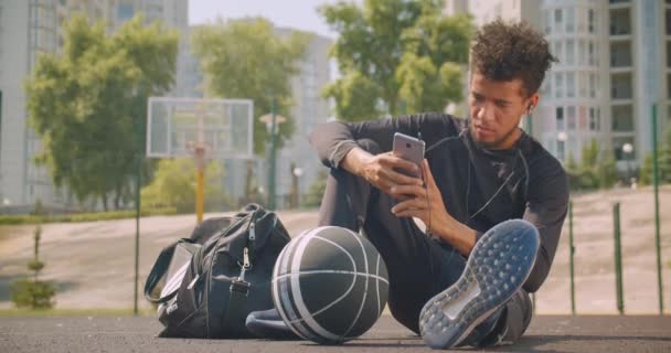 若いハンサムなアフリカ系アメリカ人男性バスケットボール選手のクローズアップ肖像画は、背景に建物とコートの屋外でボールで座って彼の携帯電話上のバイブで音楽を聴く — ストック動画