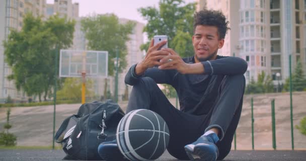 Retrato de primer plano de un joven fuerte jugador de baloncesto afroamericano tomando selfies en el teléfono sentado con una pelota al aire libre en la cancha — Vídeo de stock