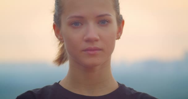Крупный план портрета молодой довольно спортивной бегуньи в черной футболке, смотрящей в камеру с красивым закатом на заднем плане — стоковое видео