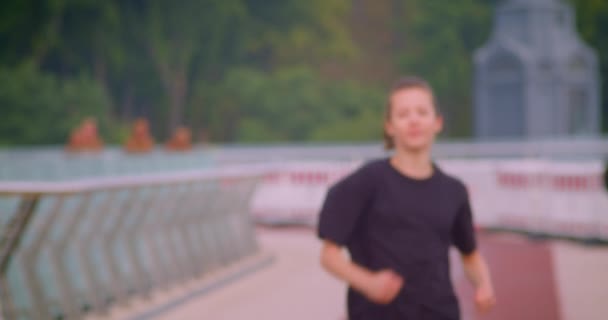 Nahaufnahme Porträt einer jungen, ziemlich sportlichen Joggerin in einem schwarzen T-Shirt, die mit verschwommenem Fokus auf der Brücke in der Großstadt im Freien läuft — Stockvideo