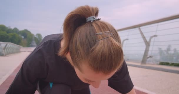 Nahaufnahme Porträt einer jungen entschlossenen, sportlichen Joggerin im schwarzen T-Shirt, die in einer Startposition sitzt und auf der Brücke in der Großstadt im Freien läuft — Stockvideo