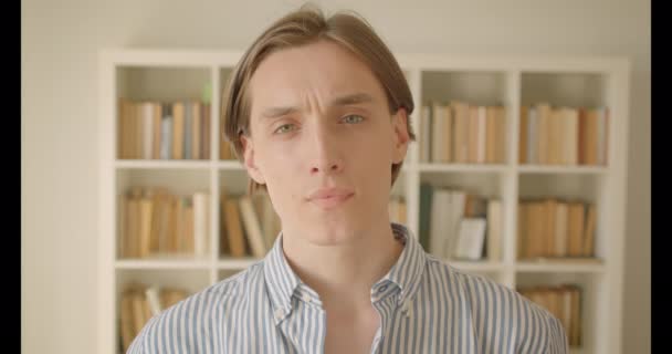 Retrato de primer plano de un joven estudiante caucásico mirando la cámara de la biblioteca en el interior con estanterías en el fondo — Vídeos de Stock