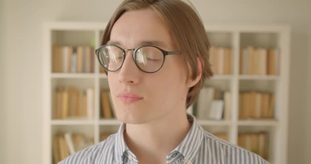 Κοντινό πλάνο του νεαρού ελκυστική Καυκάσιος αρσενικό φοιτητής σε γυαλιά κοιτάζοντας την κάμερα χαμογελά χαρούμενα στη βιβλιοθήκη σε εσωτερικούς χώρους — Αρχείο Βίντεο