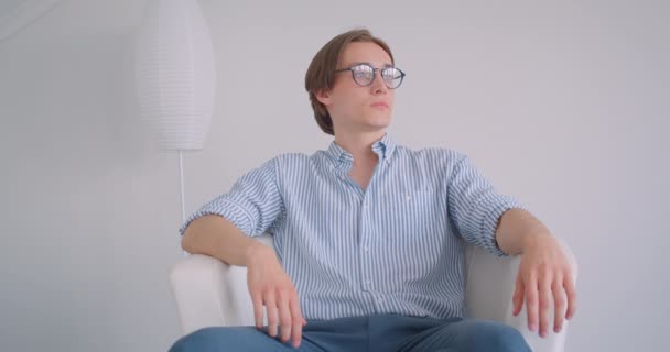 Nahaufnahme Porträt eines jungen attraktiven kaukasischen Geschäftsmannes mit Brille, der in einer weißen Wohnung im Sessel sitzt und in die Kamera blickt — Stockvideo