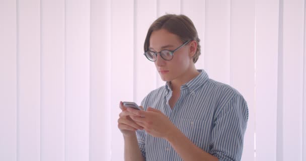 Nahaufnahme Porträt eines jungen attraktiven kaukasischen Geschäftsmannes in Brille mit dem Telefon, das drinnen im Büro steht — Stockvideo