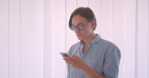 Крупный план портрета молодого привлекательного кавказского бизнесмена в очках, который пишет смс по телефону, стоя в помещении офиса — стоковое видео