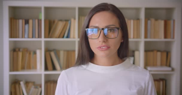 Primer plano retrato de la joven mujer de negocios bastante caucásica en gafas mirando a la cámara con estanterías en el fondo — Vídeo de stock