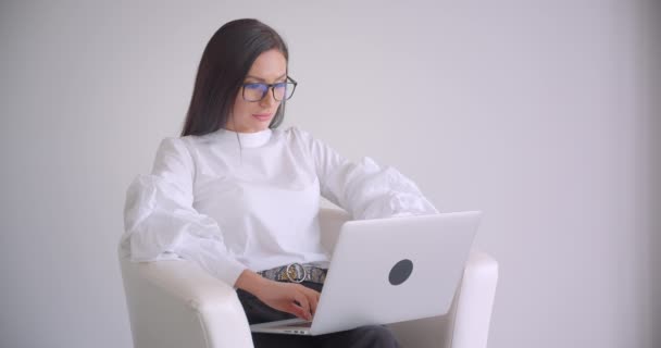Primer plano retrato de la joven mujer de negocios bastante caucásica en gafas de trabajo en el portátil sentado en el sillón con fondo blanco — Vídeos de Stock