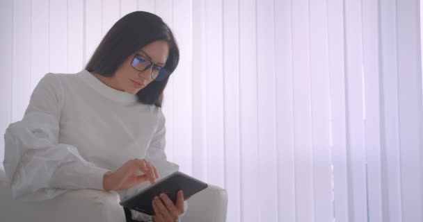 Κοντινά πλάνα πορτρέτο της νεαρής όμορφη Καυκάσιος επιχειρηματίας σε γυαλιά χρησιμοποιώντας το δισκίο κοιτάζοντας την κάμερα χαμογελώντας ευτυχισμένα κάθεται στην πολυθρόνα — Αρχείο Βίντεο