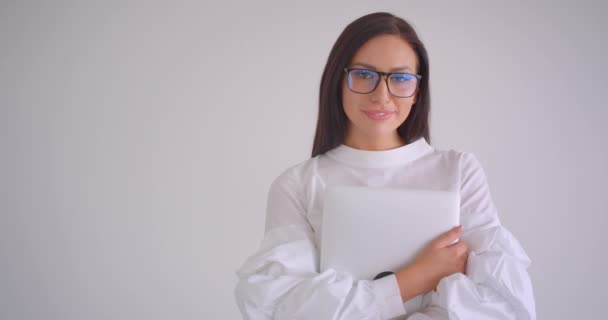 Retrato de cerca de la joven mujer de negocios bastante caucásica en gafas con un portátil mirando a la cámara de pie en la oficina blanca — Vídeos de Stock