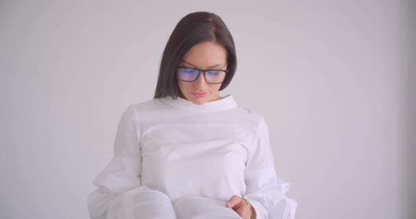 Крупный план портрета молодой симпатичной кавказской предпринимательницы в очках, читающей книгу, стоящую в белом кабинете — стоковое видео