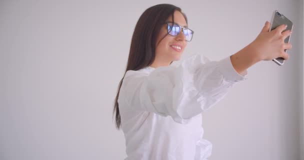 Крупный план портрета молодой симпатичной кавказской предпринимательницы в очках, делающей селфи по телефону, стоящему в белом кабинете — стоковое видео