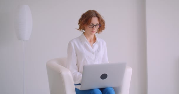 Gros plan portrait d'adulte rousse caucasien femme d'affaires dans des lunettes à l'aide de l'ordinateur portable en regardant la caméra souriant heureux assis dans le fauteuil dans le bureau blanc — Video