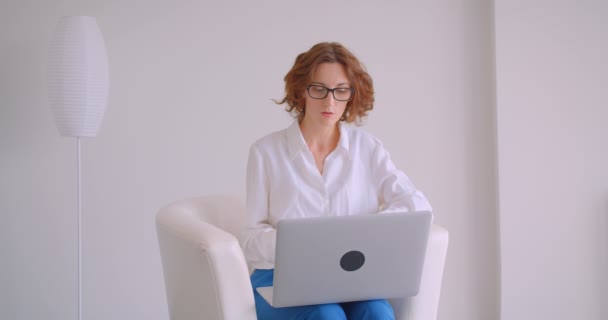 Retrato de close-up de mulher de negócios caucasiana ruiva adulta em óculos com uma chamada de vídeo no laptop sentado na poltrona no escritório branco — Vídeo de Stock