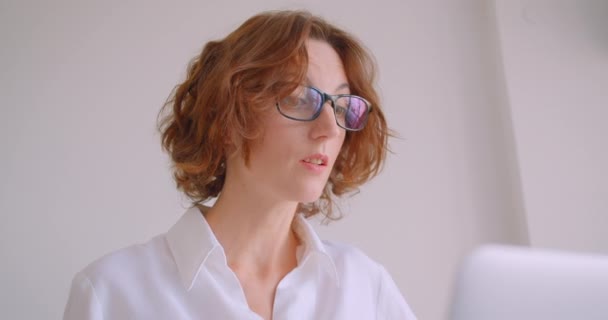 Primer plano retrato de pelirroja adulta mujer de negocios caucásica en gafas usando el portátil siendo infeliz sentado en el sillón en la oficina blanca — Vídeo de stock