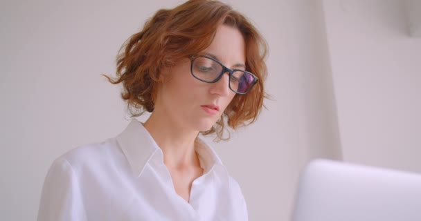 Nahaufnahme Porträt einer rothaarigen kaukasischen Geschäftsfrau mit Brille, die den Laptop benutzt und in die Kamera lächelt, glücklich im Sessel im weißen Büro sitzend — Stockvideo