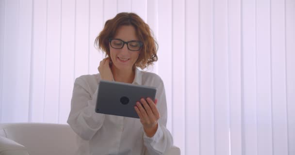Крупный план портрета взрослой рыжеволосой привлекательной бизнесвумен в очках с помощью планшета, сидящего в кресле в белом кабинете — стоковое видео