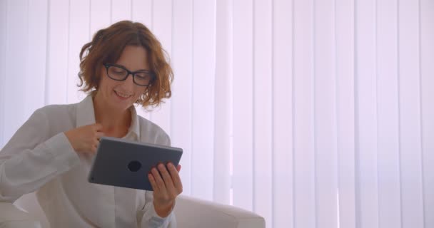 Beyaz ofiste koltukta mutlu bir şekilde oturan kameraya bakan tableti kullanarak gözlüklü yetişkin kızıl saçlı çekici iş kadınının yakın çekim portresi — Stok video
