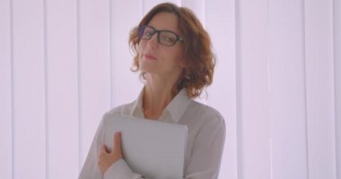 Beyaz dairede iç mekanlarda mutlu bir şekilde gülümseyen kameraya bakan bir dizüstü bilgisayar tutan gözlüklü kızıl saçlı beyaz iş kadınının yakın çekim portresi