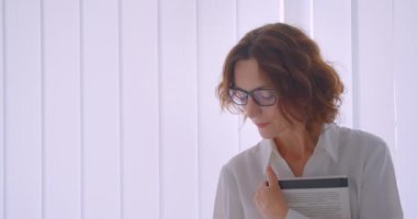 Beyaz dairede mutlu bir şekilde gülümseyen kameraya bakan bir kitap tutan gözlüklü yetişkin kızıl saçlı beyaz iş kadınının yakın çekim portresi