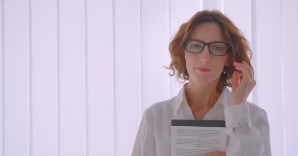 Gros plan portrait d'adulte rousse caucasien femme d'affaires dans des lunettes tenant un livre regardant la caméra à l'intérieur dans l'appartement blanc — Video