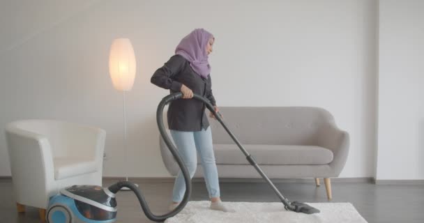 Крупный план портрета молодой мусульманки в хиджабе с помощью пылесоса и выполняя домашнюю работу в помещении квартиры — стоковое видео
