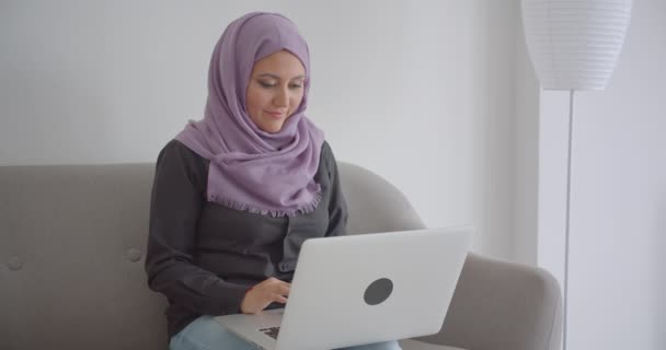 Κοντινό πλάνο της νεαρής μουσουλμανικής επιχειρηματία στη μαντίλα χρησιμοποιώντας το φορητό υπολογιστή που κάθεται στον καναπέ μέσα στο διαμέρισμα — Αρχείο Βίντεο