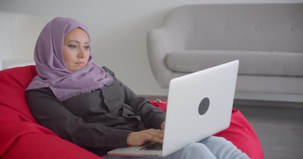 Портрет крупным планом молодой мусульманки в хиджабе с ноутбуком, сидящей в мешке, смотрящей в камеру и радостно улыбающейся в помещении — стоковое видео