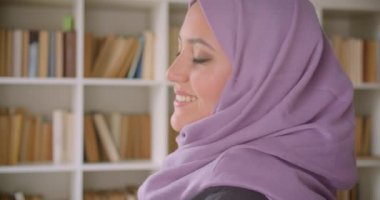 Türbanlı genç güzel müslüman kız öğrencinin yakın çekim portresi ve arka planda kitaplıklarla kütüphanede mutlu bir şekilde gülümseyen kameraya bakmak