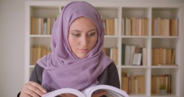Türbanlı genç, müslüman kız öğrencinin kütüphanede kameraya bakan bir kitap okumasının yakın çekim portresi