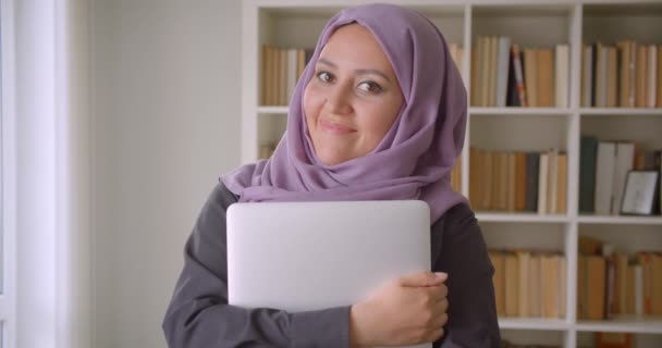 Крупним планом портрет молодої досить мусульманської студентки-жінки в Хіджабі тримає ноутбук, дивлячись на камеру, що щасливо посміхається в бібліотеці — стокове відео