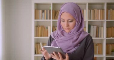 Tablet kullanarak başörtüsü genç güzel Müslüman kadın Closeup portre ve kütüphanede duran kamera bakarak