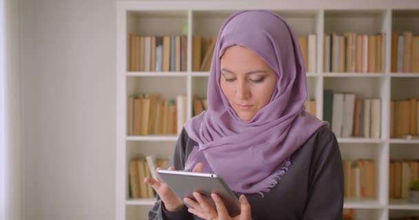 Close-up portret van jonge mooie moslim vrouw in hijab met behulp van de Tablet en kijken naar de camera staande in de bibliotheek — Stockvideo