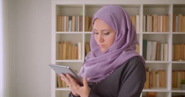 Retrato de cerca de una joven mujer bastante musulmana en hijab usando la tableta y mirando la cámara de pie en la biblioteca en el interior — Vídeo de stock