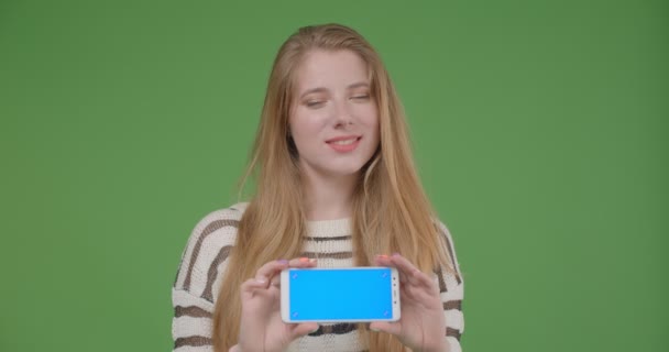 Närbild shoot av unga ganska kaukasiska hona med hjälp av mobiltelefon och visar blå skärm till kameran håller den horisontellt med isolerad bakgrund — Stockvideo