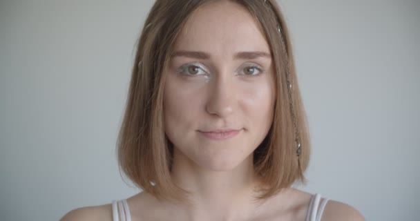 Κοντινό γύρισμα της νεαρής όμορφο καυκάσιο θηλυκό με δαχτυλίδια μαλλιών και λάμψη μακιγιάζ κοιτάζοντας την κάμερα στο λευκό δωμάτιο σε εσωτερικούς χώρους — Αρχείο Βίντεο