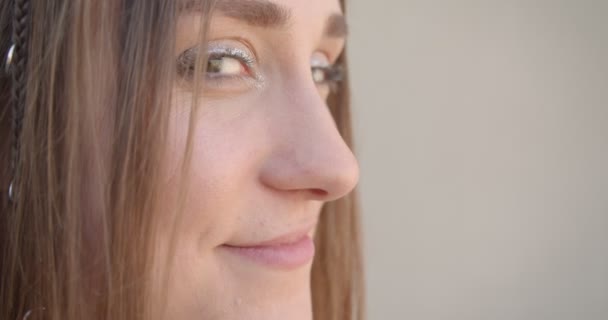 Съемка лица молодой белой женщины с кольцами для волос и блестящим макияжем с глазами, смотрящими на камеру в белом помещении — стоковое видео