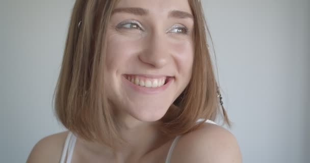 Съемки крупным планом молодой белой девушки с кольцами для волос и блестящим макияжем, счастливо смотрящей в сторону в белом помещении — стоковое видео