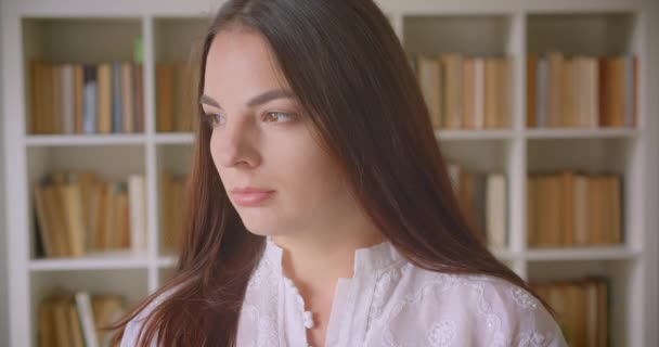 Close-up portret van jonge mooie blanke vrouwelijke student kijkend naar camera binnenshuis in de bibliotheek met boekenkasten op de achtergrond — Stockvideo
