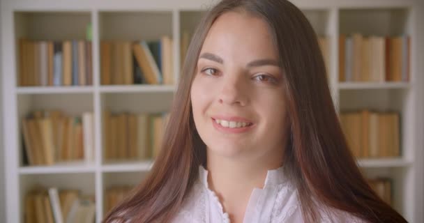 Closeup portrait of young pretty caucasian female student looking at camera smiling happily indoors in the library — Stock Video