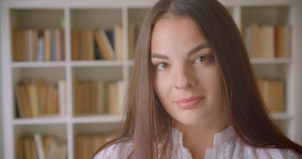 Closeup portrait of young pretty caucasian female student looking at camera smiling happily indoors in the library with bookshelves on the background — Stock Video