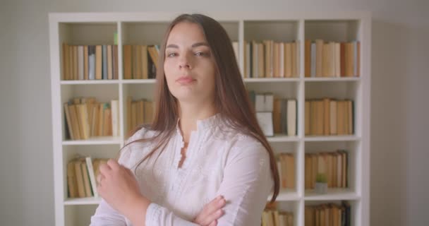 Retrato de cerca de una joven estudiante bastante caucásica mirando a la cámara con los brazos cruzados sobre el pecho dentro de la biblioteca — Vídeos de Stock