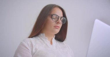 Beyaz ofiste kapalı kanepede oturan kameraya bakarak dizüstü bilgisayar üzerinde çalışan gözlüklü genç kafkas iş kadının Closeup portre