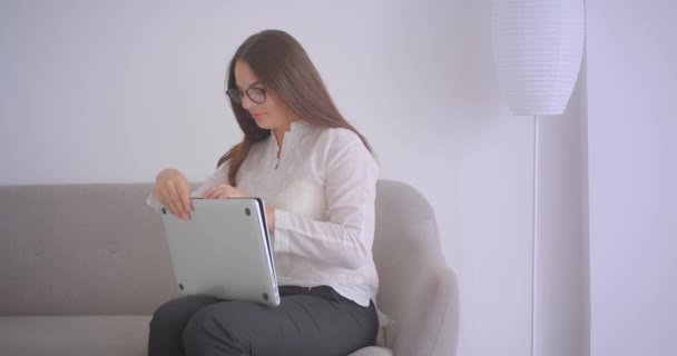 Gros plan portrait de jeune femme d'affaires caucasienne en lunettes travaillant sur l'ordinateur portable assis sur le canapé à l'intérieur dans l'appartement blanc — Video