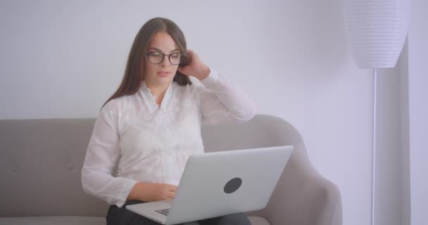 Closeup portré fiatal kaukázusi üzletasszony a szemüveg, amelynek videohívás a laptop ül a kanapén beltérben a fehér lakásban — Stock videók