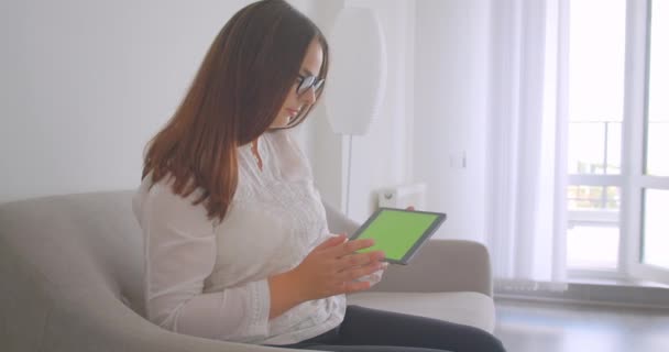 Visão lateral close-up retrato de mulher branca jovem em óculos usando o tablet e mostrando tela de croma verde para câmera sentada no sofá dentro de casa — Vídeo de Stock