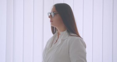Gözlüklü ve beyaz önlüklü genç beyaz kadın doktorun beyaz dairede kapalı alanda duran kameraya bakan yakın çekim portresi