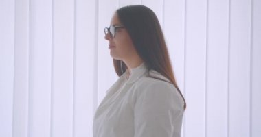 Gözlüklü ve beyaz önlüklü genç beyaz kadın doktorun beyaz odada kapalı alanda duran kameraya güvenle bakan yakın çekim portresi