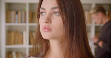 Kütüphanede duran kameraya bakan genç, beyaz beyaz kız öğrencinin arka planda kitap okuyan genç bir erkekle birlikte yakın çekim portresi